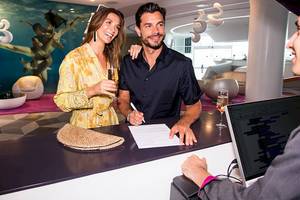 Paar im Temptation Cancun Resort Hotel, der Mann unterschreibt am Empfang, während die Frau Champagner hält.