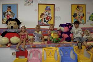 Vier Kinder sitzen auf bunten Stühlen neben großen Plüschtieren und drei Clown-Gemälden an der Wand.