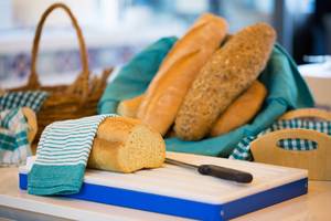 Ein Brot auf einem blauen Schneidebrett mit einem Korb voller Brötchen im Hintergrund.