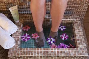 Füße in einem quadratischen Wasserbecken mit Blumen, Handtuch und Ahava-Produkt.