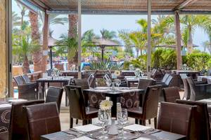 Innenansicht des Restaurants im Barcelo Fuerteventura Mar Hotel mit Tisch, Stühlen und Palmen im Hintergrund.