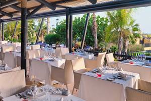 Außenbereich des Restaurants im Barcelo Fuerteventura Mar Hotel mit gedeckten Tischen und Stühlen, umgeben von Palmen und Blick auf den Garten.