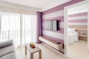 Innenansicht einer Hotelsuite im Barcelo Fuerteventura Mar mit lila und weißen Wänden, einem Sofa, einem Tisch und einem Fernseher.