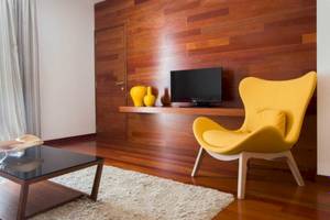 Ein Wohnzimmer mit einer holzverkleideten Wand, einem gelben Stuhl, einem Glastisch und einem Fernseher auf einem Regal.