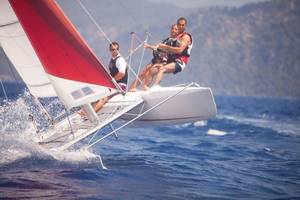 Ein Segelboot mit drei Leuten, zwei sitzen auf dem Kiel und einer vorne, im Wasser, Hillside Beach Club