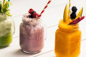 Drei Smoothie-Gläser mit verschiedenen Früchten und Strohhalmen auf einem weißen Tisch.