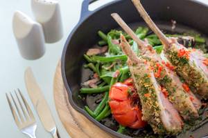 Ein Gericht in einer Pfanne mit gegrillten Lammkoteletts und Gemüse, einschließlich grüner Bohnen und Tomaten, serviert auf einem Holzuntersetzer mit Messer, Gabel und Salz- und Pfefferstreuer.