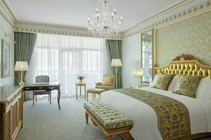 Suite-Schlafzimmer im Raffles The Palm Dubai mit großem Bett, goldenem Kopfteil, Kristallkronleuchter, Schreibtisch und Sesseln.