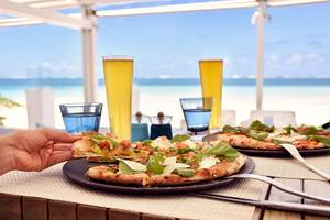 Eine Person nimmt ein Stück Pizza vom Teller mit Meer im Hintergrund.