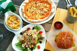 Verschiedene Teller mit Essen auf einem Holztisch, einschließlich einer Pizza, einem Salat, einem Burger und frittierten Pommes.