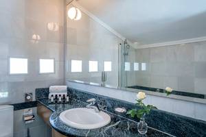 Badezimmer im Mitsis Lindos Memories Resort & Spa mit weißem Waschbecken, Marmorboden und großen Spiegel.