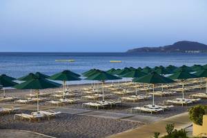 Foto der Strandseite des Esperos Village Blue & Spa Hotels mit vielen Sonnenliegen und grünen Sonnenschirmen.
