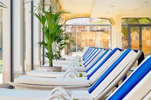 Reihe von weißen und blauen Sonnenliegen im Innenbereich des Secrets Bahia Real Resort & Spa mit Pflanzen und Glasfenstern im Hintergrund.