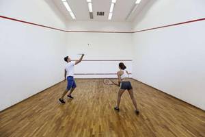 Zwei Personen spielen Squash in einer Sporthalle mit Holzboden und roten Linien.