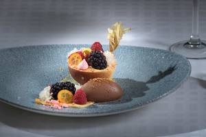 Ein reichhaltiges Dessert mit Früchten, Schokolade und Beilagen auf einem blauen Teller serviert.