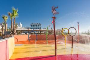 Wasserspielplatz im Freien mit bunten Wassersprudlern und Gebäuden im Hintergrund.