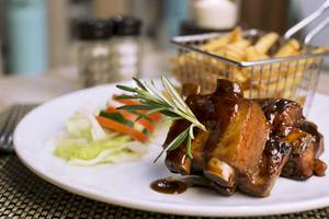 Ein Teller mit gebratenen Schweinerippchen, Salat und Pommes Frites im Hintergrund.