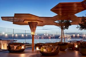 Myconian Naia Hotel Terrasse bei Nacht mit Blick auf den Ozean und einer beleuchteten Stadt im Hintergrund.