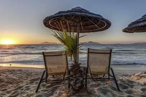 Sonnenuntergang am Strand mit zwei Liegen und einer Palme im Vordergrund.