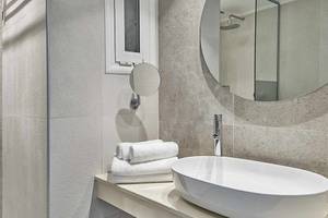 Badezimmer im Atlantica Marmari Palace mit grauen Fliesen, einem runden Spiegel und einem Waschbecken.