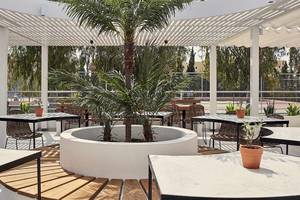 Im Freien gelegenes Restaurant des Atlantica Marmari Palace Hotels mit Tischen, Stühlen und einer großen Palme in der Mitte.