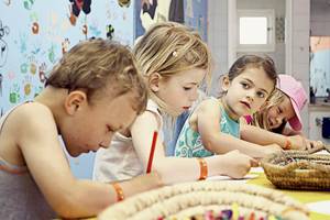 Vier Kinder sitzen an einem Tisch und zeichnen, ein Korb mit Buntstiften steht vor ihnen.