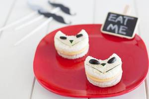 Zwei Katzen-Sandwiches auf einem roten Teller mit einem schwarzen Schild, das 'Eat Me' sagt, und Mustache-Picks im Hintergrund.