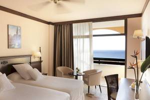 Doppelzimmer mit Meerblick im Occidental Jandia Playa Hotel, mit zwei Betten, Sesseln, einem Tisch und einem großen Fenster mit Balkon.