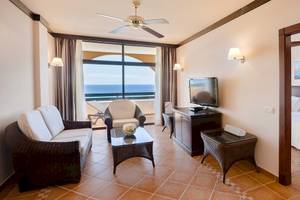 Hotelzimmer mit Meerblick, Wohnzimmerbereich mit Sofa, Tisch, Fernseher und Balkon.