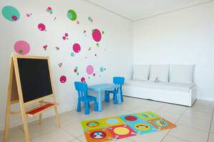 Ein Kinderzimmer mit einem blauen Spieltisch, zwei blauen Stühlen, einer Bank, einem Teppich und einer Tafel.