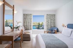 Innenansicht eines Schlafzimmers im Mitsis Ramira Beach Hotel mit Bett, Schreibtisch, Spiegel und Balkonzugang.