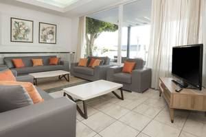 Wohnbereich des Kosta Palace City Hotels mit grauen Sofas, orangen Kissen und einer großen Fensterfront.