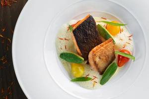 Ein Fischgericht in einem weißen Teller auf einem Holztisch mit verschiedenen Gemüsen und Gewürzen.