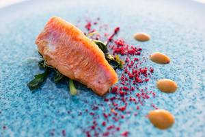 Gebratener Fisch auf blauem Teller mit grünem Blattwerk und roten und gelben Toppings.