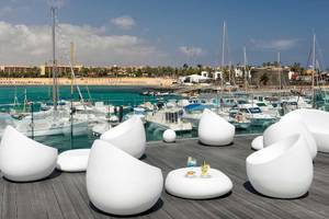 Terrasse des Barcelo Fuerteventura Castillo mit weißen Stühlen und Tischen, Blick auf den Hafen mit vielen Booten.