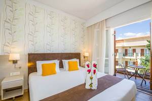 Zimmer im Kalyves Beach Hotel mit Balkon, Bett, Nachttischen und Ausblick auf eine Terrasse.