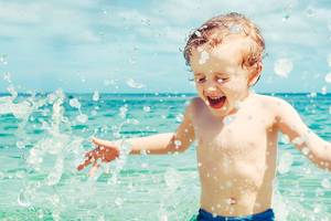 Kleiner Junge, der mit Wasser spielt und lacht, während er im Ozean steht.
