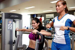 Frau im Fitnessstudio trainiert mit einer Trainerin auf einer Brustpresse-Maschine.