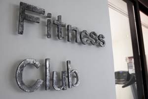 Schild mit den Worten 'Fitness Club' an einer Wand, mit einem Blick auf eine Tretmühle im Hintergrund.