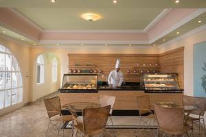 Innenansicht des Restaurants des Mitsis Family Village Beach Hotels mit einem Koch am Tresen, umgeben von Tisch und Stühlen.