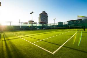 Tennisplatz mit Zäunen und Gebäuden im Hintergrund, alles in hellem Sonnenlicht
