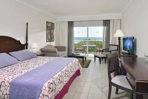 Hotelzimmer mit Meerblick und Balkon, Bett, Stühle, Schreibtisch und Fernseher.