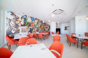 Innenbereich des Ramla Bay Resorts mit orangefarbenen Stühlen und Tischen, einer großen Wandmalerei mit Essensmotiven und modernen Kronleuchtern.