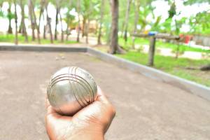 Hand hält einen silbernen Petanque-Ball vor einem Spielfeld mit Bäumen im Hintergrund.