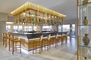 Bar im Astir Beach Hotel auf Kreta mit einer Reihe von Barhockern und einer Theke mit einer Glasvitrine oben.