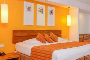 Innenansicht eines Hotelzimmers im Adaaran Select Hudhuran Fushi mit orangefarbenen Wänden, einem großen Bett, Kissen und einem Balkon mit Möbeln.