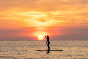 Eine Frau steht auf einem Stand-Up-Paddler bei Sonnenuntergang am Strand