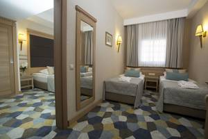 Innenansicht eines Doppelzimmers im Fame Residence Lara & Spa Hotel mit drei Betten, einem Spiegel und einem Tisch mit Blumen.