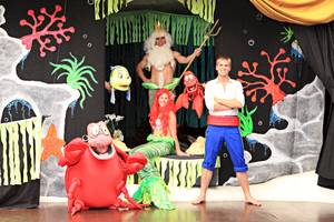 Vier Personen in Kostümen auf einer Bühne, zwei als Arielle und Sebastian, einer als König Triton und einer als Mann in einem weißen Hemd.