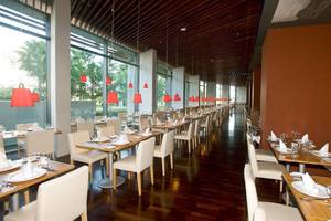 Innenansicht des Restaurants im Nirvana Cosmopolitan Hotel mit langen Reihen von Tischen und Stühlen, roten Lampen und großen Fenstern.